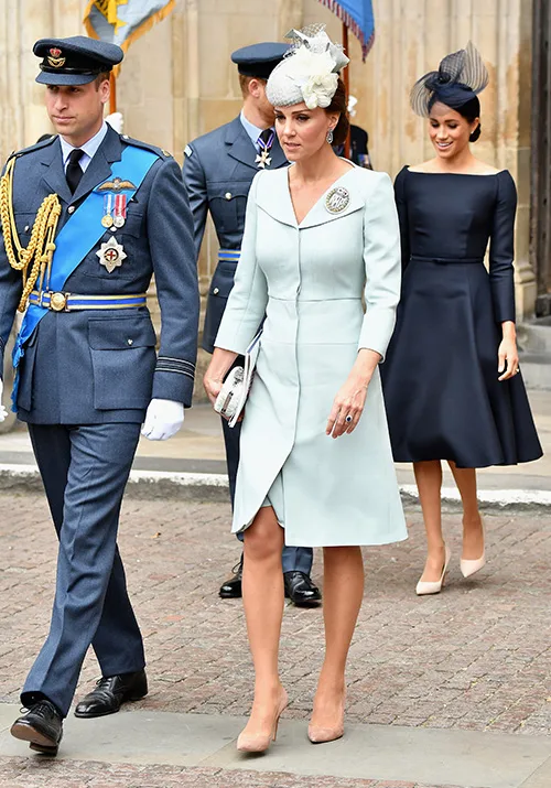 A man in military attire, a woman in a light coat dress with a hat, and another in a dark dress walk together outdoors.