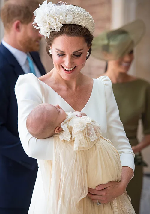 A woman in a white dress holding a baby in a christening gown, with two blurred figures in the background.
