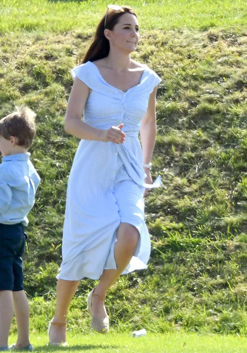 A woman in a white dress walking on grass beside a child, with her hair down and sunglasses on her head.