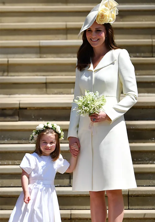 A woman in a light outfit with a matching hat stands on steps holding hands with a young girl in a white dress and flower crown.