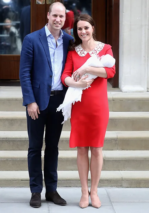 Couple stands on steps, woman in red dress holding a baby wrapped in white, posing and smiling for a photo.