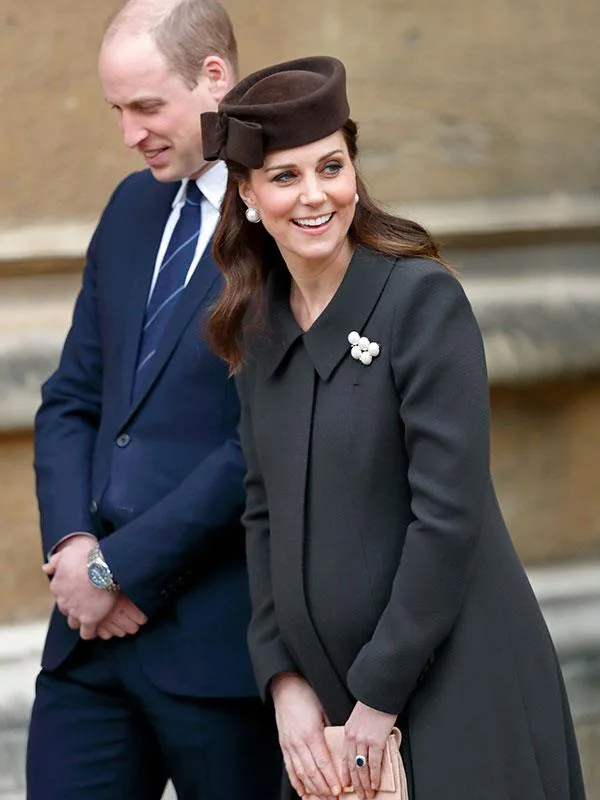 A man in a suit stands next to a smiling woman in a dark coat and hat, outdoors.