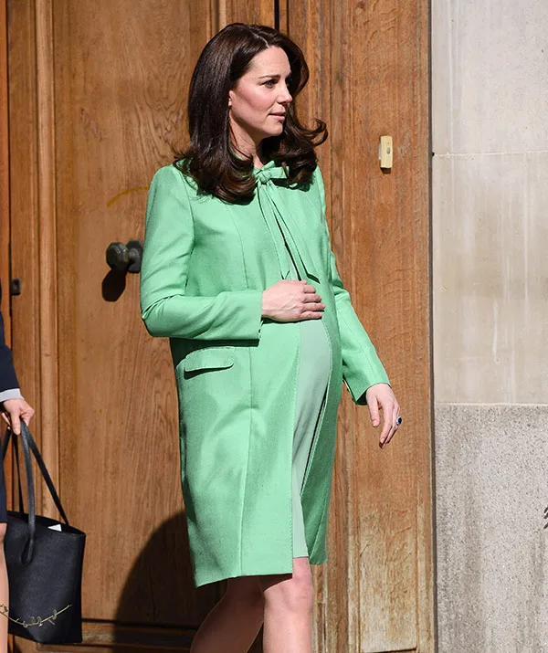 A woman in a green coat with long dark hair holds her belly, standing outside by a wooden door.