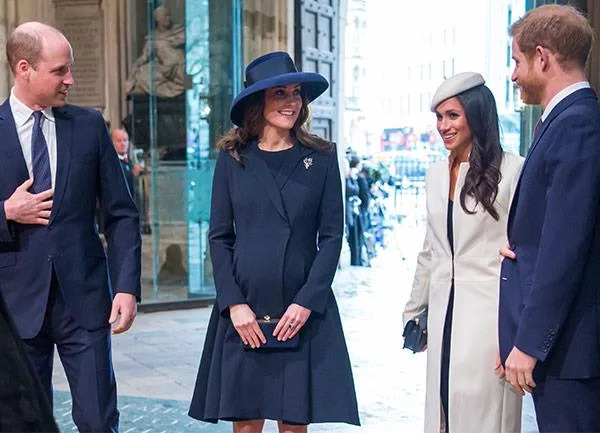 People in formal attire standing outside a building, two women in hats and men wearing suits, smiling and conversing.