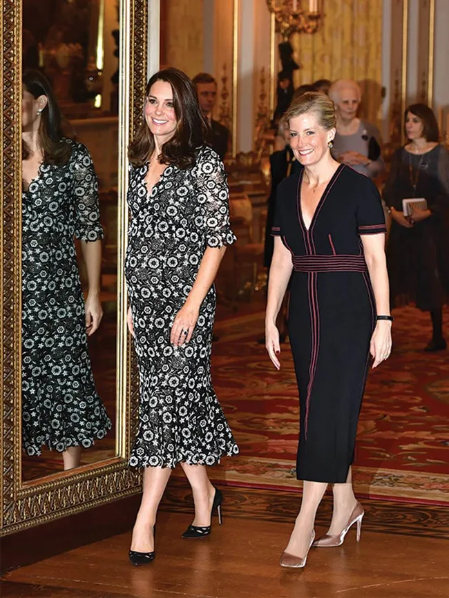 Two women in elegant dresses walk near a large ornate mirror in a grand room.