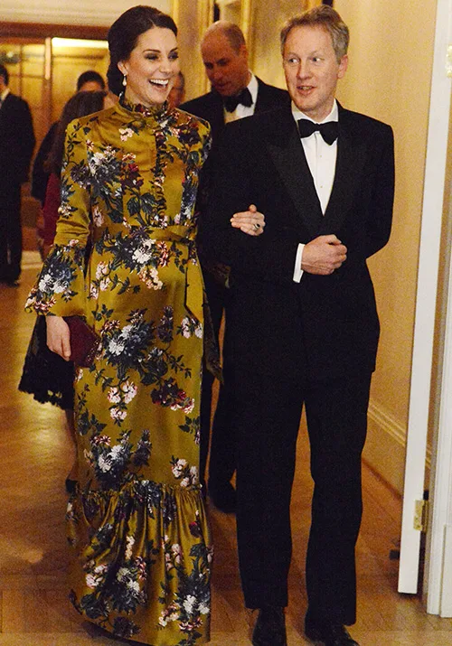 A woman in a floral dress and a man in a tuxedo walk arm-in-arm at a formal event on a wooden-floored hallway.