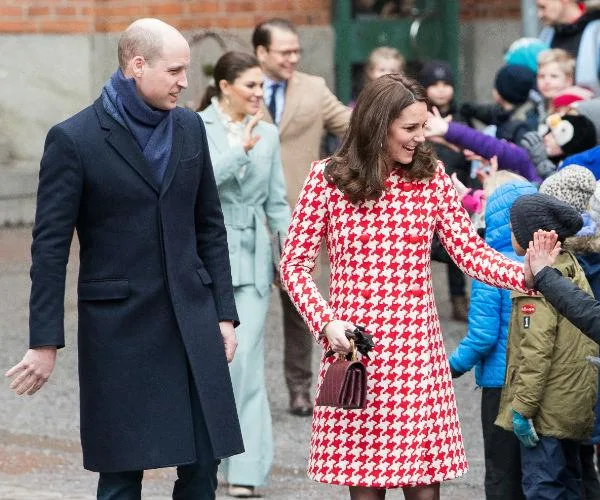 Royal couple greeting children outdoors, man in dark coat and woman in red houndstooth coat smiling and waving.