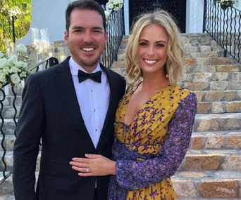 A smiling couple posing outdoors; he's in a tuxedo, she's in a floral dress, standing on stairs with flowers around.