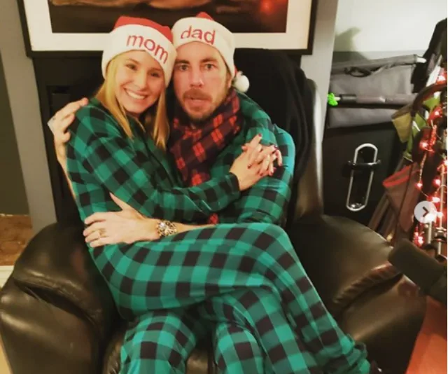 A couple in matching green plaid pajamas and "mom" and "dad" hats sitting on a chair, with holiday decor around.