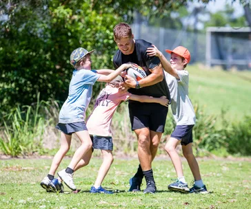 Man playing rugby with three kids in a park, all laughing and engaged in a fun, playful tackle.