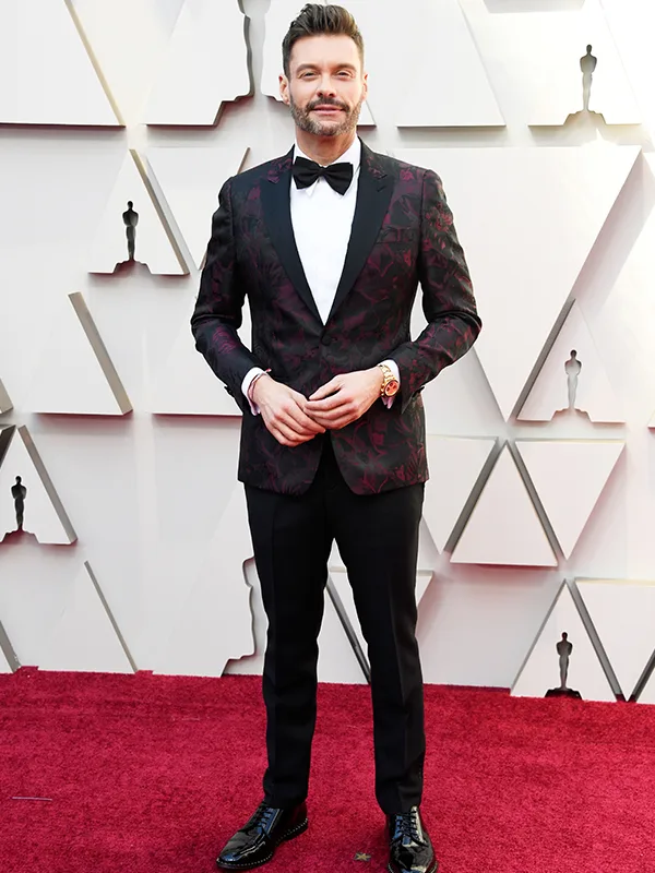 Man in a patterned suit and bow tie on the Oscars red carpet. Triangular backdrop with Oscar statuette motifs.