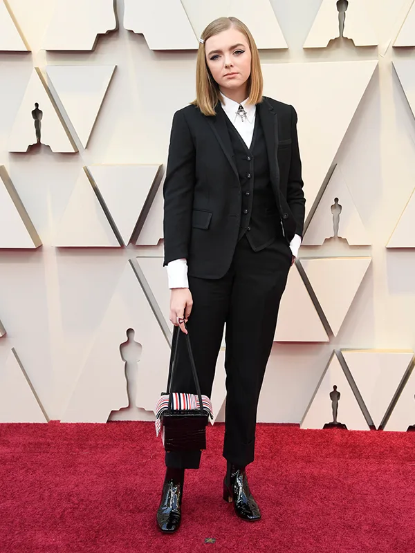Person in a black suit on a red carpet, holding a handbag, standing in front of geometric wall design.