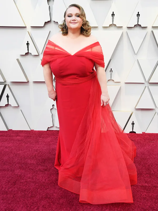 A woman in a red gown stands on a red carpet at an event with a geometric white background.