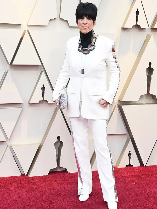 Person in a white suit on the red carpet with a geometric backdrop, holding a glittery clutch.