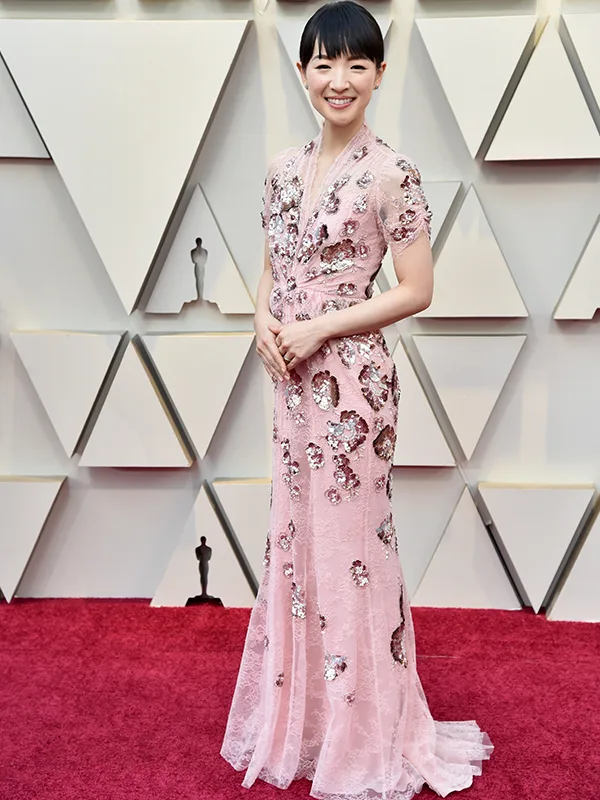 A person in a shimmering pink gown stands on a red carpet in front of a geometric-patterned wall.