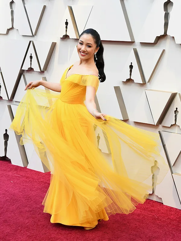 Woman in a flowing yellow gown poses on a red carpet event with geometric wall backdrop.
