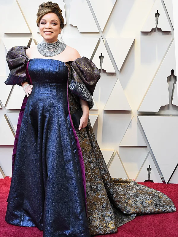A woman in a blue and gold gown on a red carpet, standing against a backdrop with geometric patterns.