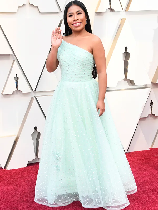 Person in a light blue, one-shoulder gown waves on a red carpet, with an Oscar-themed backdrop behind.