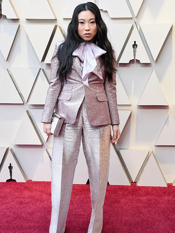Individual in a shimmering pink suit with a large bow tie, posing on a red carpet at a formal event.