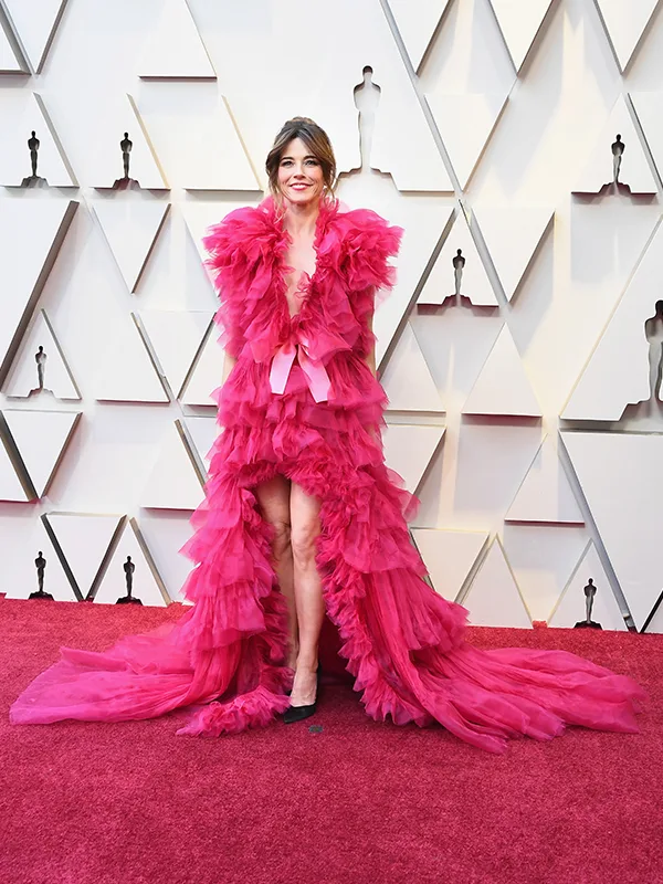 Person in a vibrant pink ruffled gown with a long train, posing on a red carpet at a formal event.