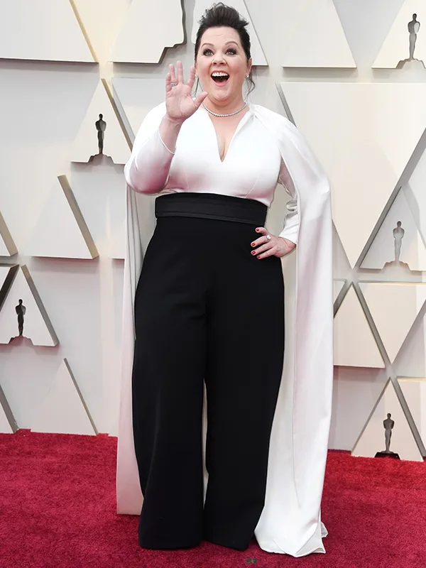 Person in a white cape and black pants waving on a red carpet with a geometric backdrop.