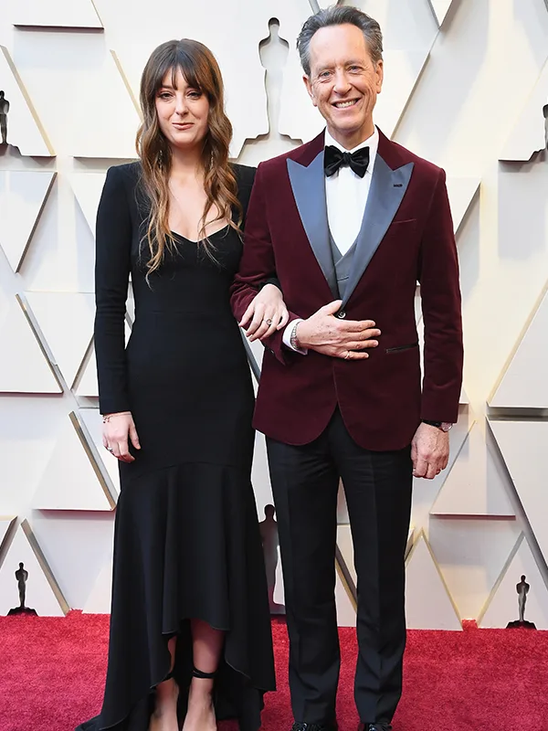 A woman in a black dress and a man in a burgundy suit pose on a red carpet with geometric background.