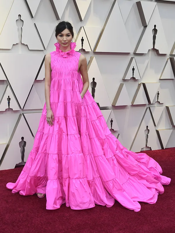 Person in pink ruffled gown poses on red carpet with geometric background.