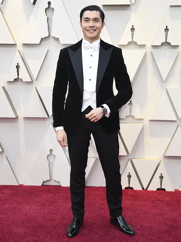 A man in a black tuxedo with a white bow tie stands on the red carpet at an awards event.