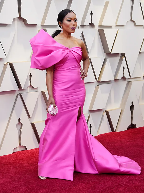 Woman in a vibrant pink gown on a red carpet at an event, standing confidently with hands on hips.