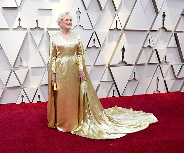 Woman in a shimmering gold gown with a long train stands on a red carpet at an event.