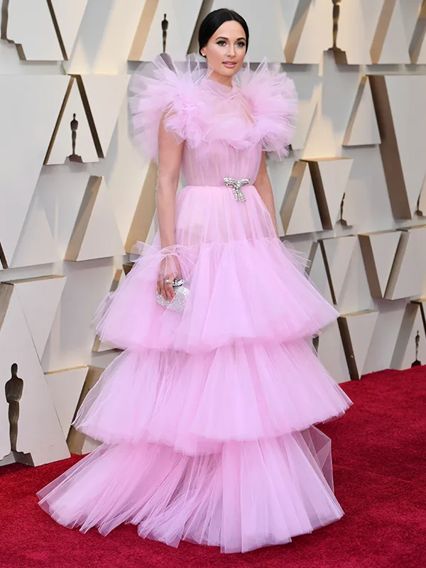 Woman in tiered pink tulle gown with ruffled shoulders, holding a silver clutch, standing on a red carpet.