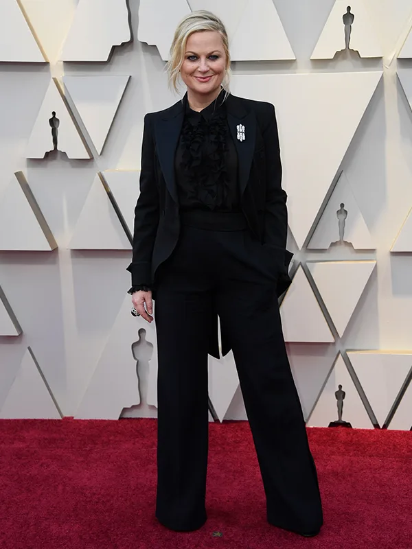 A person in a black suit with hands in pockets poses on a red carpet with geometric wall backdrop.