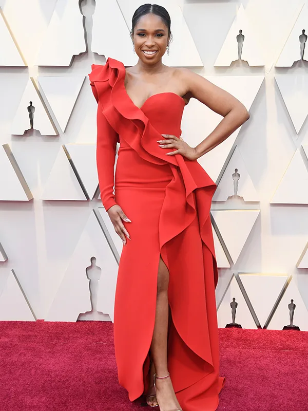 Woman in a ruffled red gown with a slit, standing on a red carpet against a geometric backdrop.