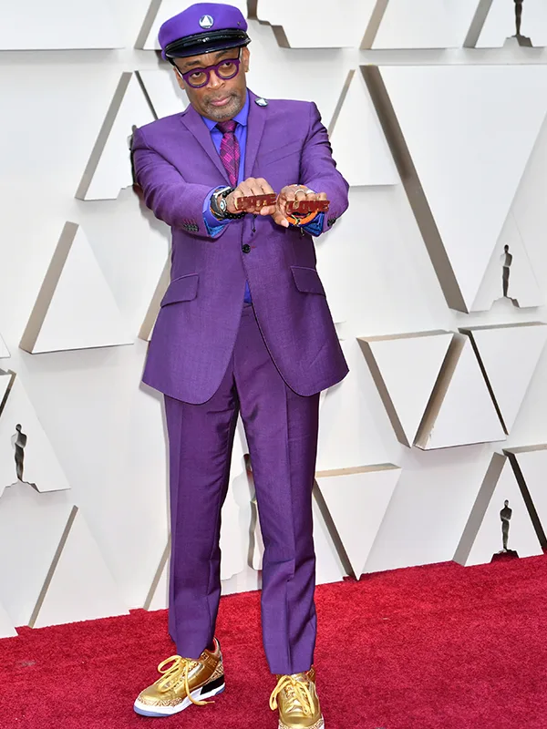 A person in a purple suit and cap on a red carpet, showing knuckles adorned with rings, wearing gold sneakers.