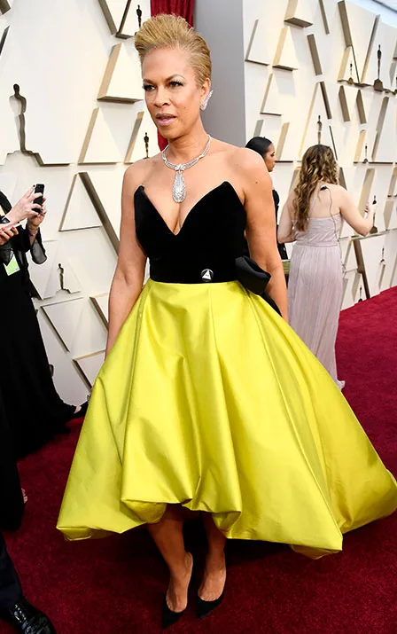 A person in a black and yellow gown on the Oscars red carpet, standing on a geometric pattern backdrop.