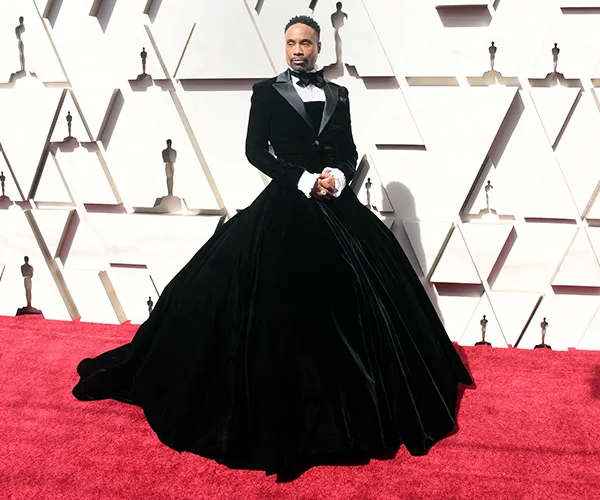 Man in a velvet tuxedo gown on a red carpet at an event with geometric wall pattern in the background.