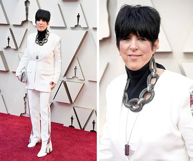 Woman in a white suit with large necklace on red carpet at award event, geometric backdrop visible.