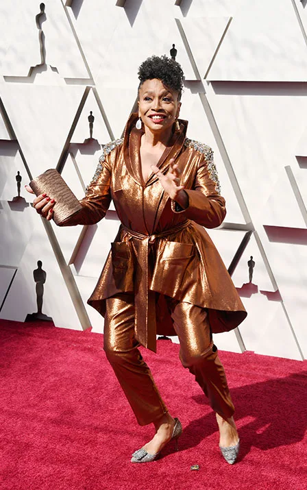 A person in a metallic bronze outfit smiles and poses on a red carpet in front of an Oscars-themed backdrop.