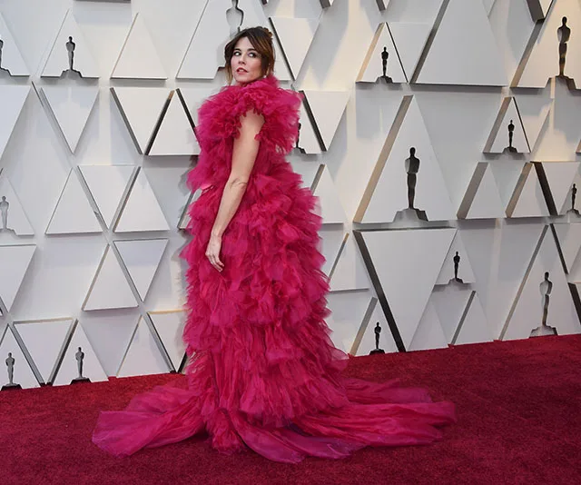 A person in a bright red ruffled gown poses on a red carpet in front of wall with triangular patterns.
