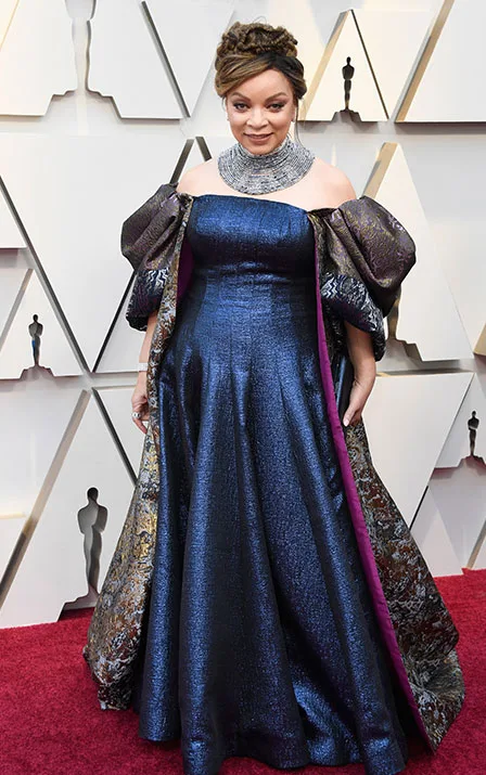 A person in an off-the-shoulder blue gown with a metallic necklace on a red carpet, with geometric background patterns.