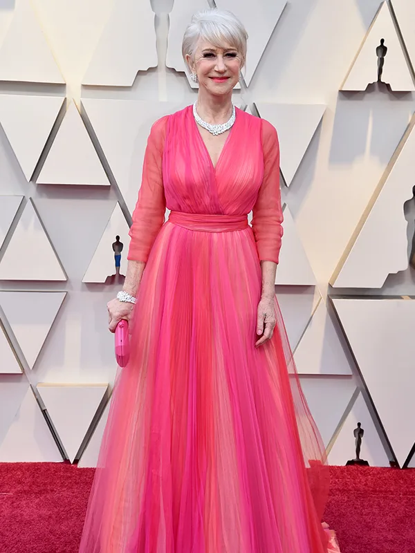Woman in a pink gown with silver jewelry poses on a red carpet with a geometric backdrop.