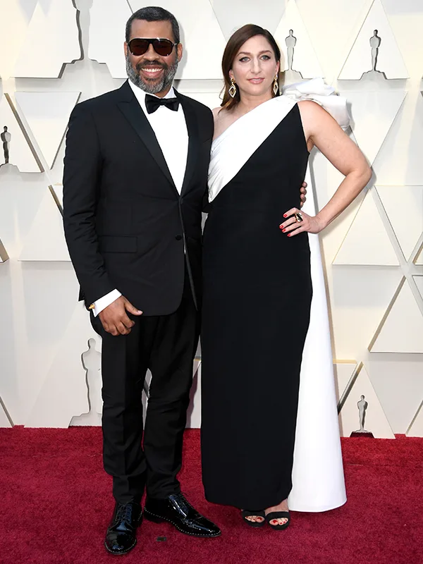 A couple on the red carpet, man in a black tuxedo and woman in a black and white gown, smile for the cameras.