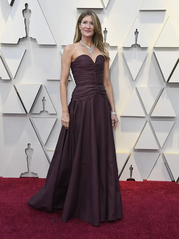 Woman in a strapless burgundy gown with a diamond necklace, posing on a red carpet in front of an Oscar-themed backdrop.