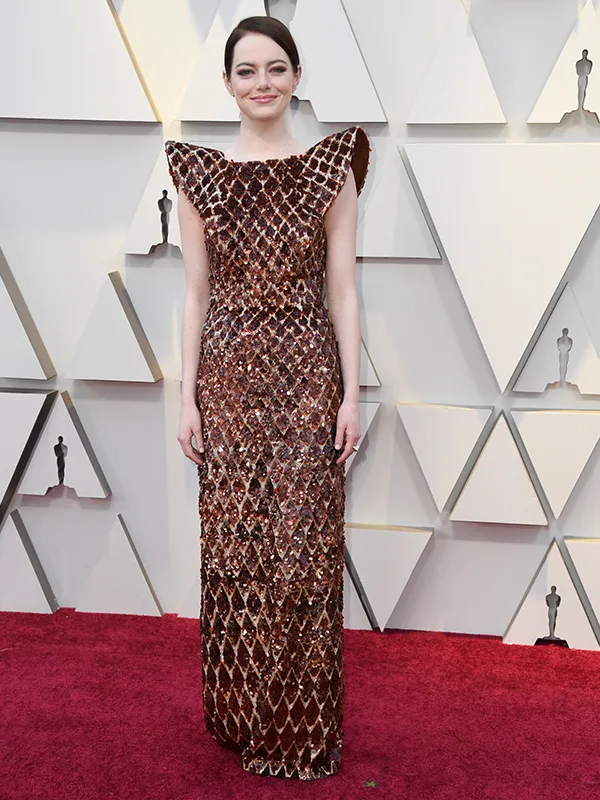 A woman in a shimmering brown dress poses on a red carpet in front of a geometric patterned backdrop.