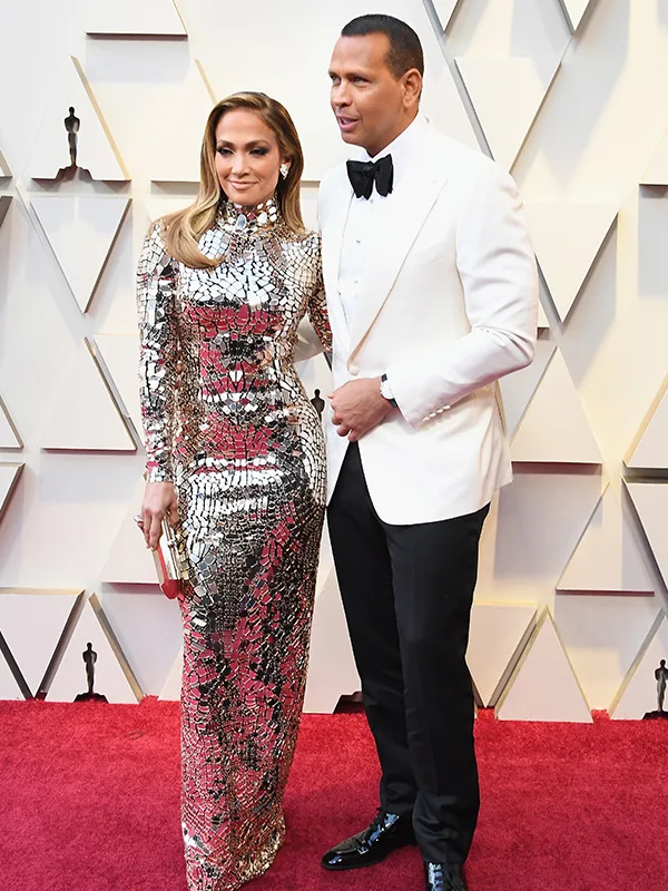 A couple poses on the red carpet; woman in a silver gown and man in a white tuxedo with a black bow tie.