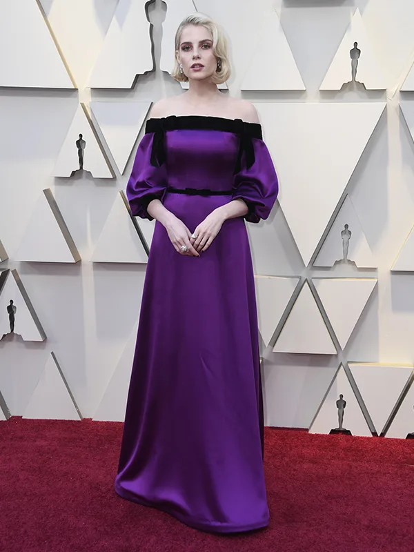 Person in a purple off-shoulder gown on a red carpet with a geometric wall backdrop.