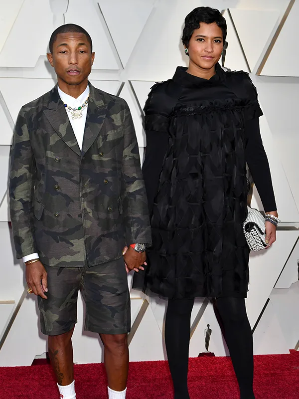 Couple holding hands on red carpet; man in camo suit and shorts, woman in black dress.