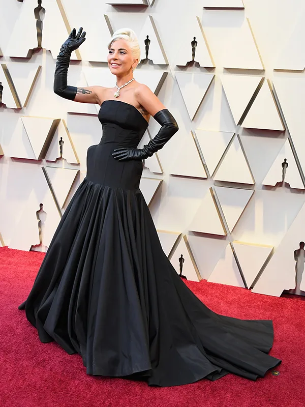 Person in elegant black gown with matching gloves waves on red carpet, geometric backdrop behind.