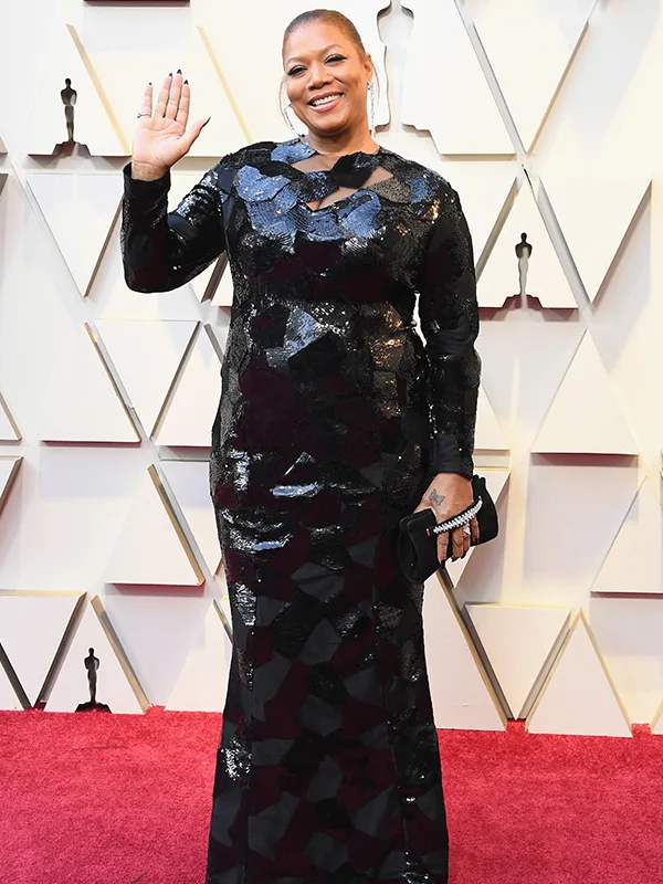 An individual in a black sequin gown waves on a red carpet at an awards event.