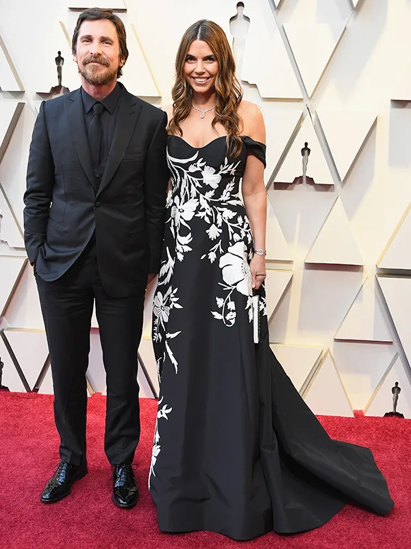 Man in a black suit and woman in a black dress with white floral pattern on a red carpet.